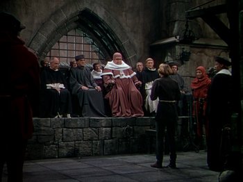 Movie still from “Joan of Arc” (1948), directed by Victor Fleming – A group of people sitting on a stone wall in front of a building; Wide shot, High angle