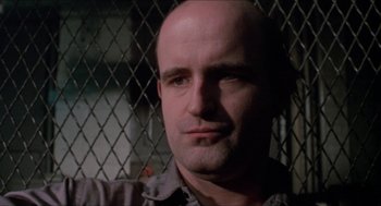 Movie still from “Joe” (1970), directed by John G. Avildsen – A man with a bald head is standing in front of a chain link fence; Close Up shot, Low angle