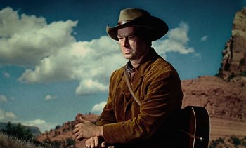 Movie still from “Johnny Guitar” (1954), directed by Nicholas Ray – A man wearing a cowboy hat sitting in a chair; Medium shot, Low angle