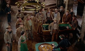 Movie still from “Johnny Guitar” (1954), directed by Nicholas Ray – A group of men and a woman standing in front of a roulette wheel; Wide shot, High angle