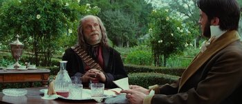 Movie still from “Jonah Hex” (2010), directed by Jimmy Hayward – An older man sitting at a table with a glass of water; Medium shot, Over the shoulder angle