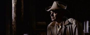 Movie still from “Jubal” (1956), directed by Delmer Daves – A man wearing a cowboy hat in a dimly - lit room; Close Up shot, Over the shoulder angle