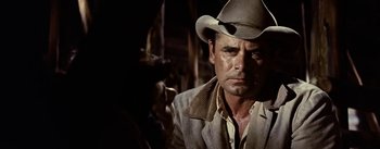 Movie still from “Jubal” (1956), directed by Delmer Daves – A man wearing a cowboy hat in a dimly - lit room; Close Up shot, Over the shoulder angle