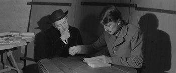Movie still from “Jules and Jim” (1962), directed by François Truffaut – Two men sitting at a wooden table and one is reading a book; Medium shot, Over the shoulder angle