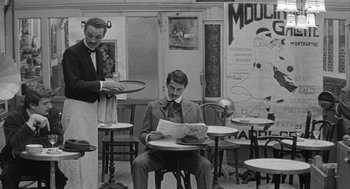 Movie still from “Jules and Jim” (1962), directed by François Truffaut – A man sitting at a table reading a newspaper while another man holds a plate; Wide shot, Over the shoulder angle