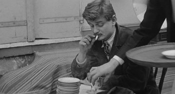 Movie still from “Jules and Jim” (1962), directed by François Truffaut – A man smoking a cigarette while sitting at a table with plates; Medium shot, High angle