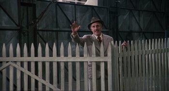 Movie still from “Julia” (1977), directed by Fred Zinnemann – A man in a suit and hat standing next to a white picket fence; Wide shot, Low angle