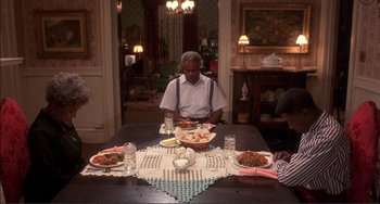 Movie still from “Jungle Fever” (1991), directed by Spike Lee – A man sitting at a dinner table with plates of food on it; Wide shot, High angle