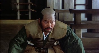 Movie still from “Kagemusha” (1980), directed by Akira Kurosawa – A man with a bald head sitting in a chair; Close Up shot, Low angle