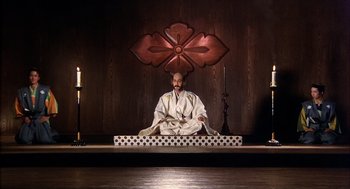 Movie still from “Kagemusha” (1980), directed by Akira Kurosawa – A man sitting on a stage in a white robe; Wide shot, Low angle