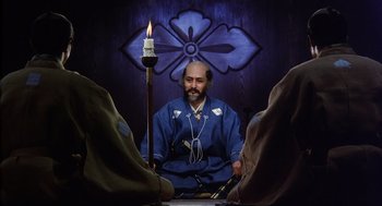 Movie still from “Kagemusha” (1980), directed by Akira Kurosawa – A man sitting in front of a candle in a room; Medium shot, Low angle
