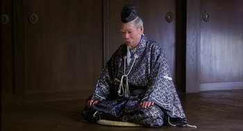 Movie still from “Kagemusha” (1980), directed by Akira Kurosawa – An older man sitting on the ground wearing a hat; Medium shot, High angle