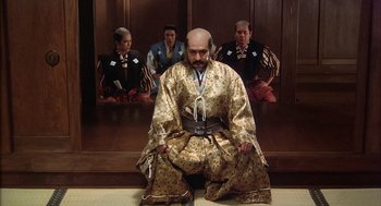 Movie still from “Kagemusha” (1980), directed by Akira Kurosawa – A man sitting on the ground in front of a group of people; Wide shot, Low angle