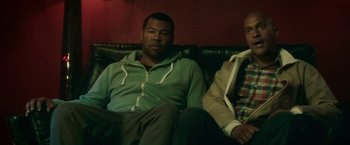Movie still from “Keanu” (2016), directed by Peter Atencio – Two men sitting on a couch in front of a red wall; Medium shot, Over the shoulder angle