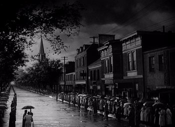 Movie still from “Keeper of the Flame” (1942), directed by George Cukor – A black and white photo of people walking down the street with umbrellas; Extreme Wide shot, High angle
