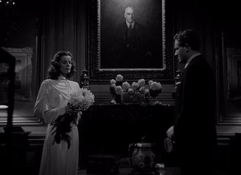 Movie still from “Keeper of the Flame” (1942), directed by George Cukor – A man and a woman holding flowers in front of a painting; Medium shot, Low angle