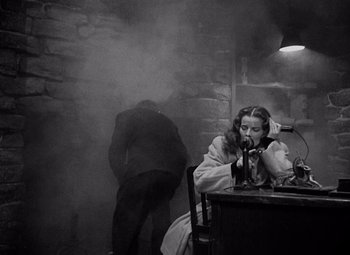 Movie still from “Keeper of the Flame” (1942), directed by George Cukor – A woman sitting at a table in a dark room; Medium shot, Low angle