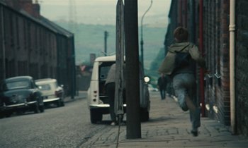 Movie still from “Kes” (1969), directed by Ken Loach – A man running down a street next to a van; Wide shot, High angle
