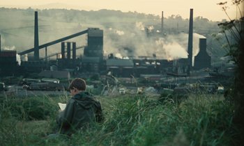 Movie still from “Kes” (1969), directed by Ken Loach – A man sitting in a field looking at a factory; Wide shot, High angle