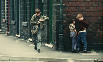 Movie still from “Kes” (1969), directed by Ken Loach – A boy walking down the street while two other boys sit on the sidewalk; Wide shot, High angle