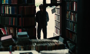 Movie still from “Kes” (1969), directed by Ken Loach – A man standing in front of shelves of books in a library; Wide shot, High angle