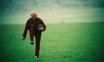 Movie still from “Kes” (1969), directed by Ken Loach – An older man in a red suit is kicking a ball; Wide shot, Low angle