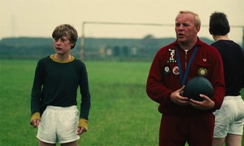 Movie still from “Kes” (1969), directed by Ken Loach – A man standing next to a young boy holding a ball; Medium shot, Over the shoulder angle