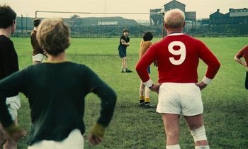 Movie still from “Kes” (1969), directed by Ken Loach – A group of young men standing on top of a lush green field; Wide shot, Over the shoulder angle