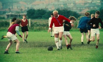 Movie still from “Kes” (1969), directed by Ken Loach – An older man kicking a soccer ball on a field; Wide shot, High angle