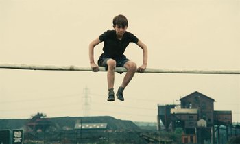 Movie still from “Kes” (1969), directed by Ken Loach – A young man sitting on top of a metal pole; Wide shot, High angle
