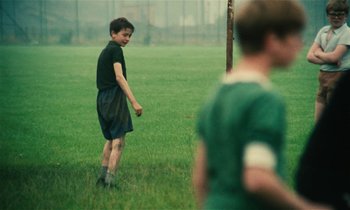 Movie still from “Kes” (1969), directed by Ken Loach – A young boy standing in a grassy field with a frisbee; Wide shot, Over the shoulder angle