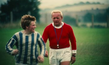 Movie still from “Kes” (1969), directed by Ken Loach – A man and a young boy walking on a grass field; Medium shot, Over the shoulder angle