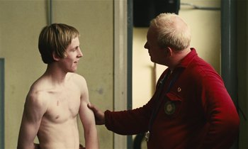 Movie still from “Kes” (1969), directed by Ken Loach – A man in a red jacket is talking to a young man; Medium shot, Over the shoulder angle