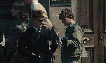 Movie still from “Kes” (1969), directed by Ken Loach – A boy holding an owl in his hand while a boy in a suit smokes a cigarette; Medium shot, Over the shoulder angle