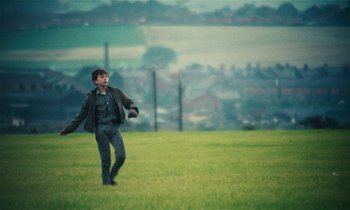 Movie still from “Kes” (1969), directed by Ken Loach – A young man is playing frisbee in a grassy field; Wide shot, Over the shoulder angle