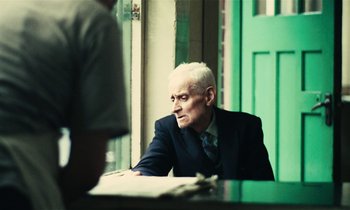 Movie still from “Kes” (1969), directed by Ken Loach – An older man sitting at a table in front of a green door; Medium shot, Over the shoulder angle