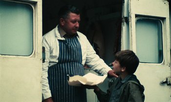 Movie still from “Kes” (1969), directed by Ken Loach – A man in an apron is handing a paper to a young boy; Medium shot, Over the shoulder angle