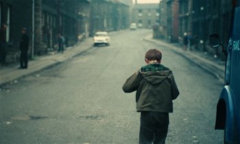 Movie still from “Kes” (1969), directed by Ken Loach – A young boy walking down the street with a car in the background; Wide shot, High angle