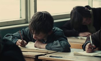 Movie still from “Kes” (1969), directed by Ken Loach – A young man writing on a piece of paper in a classroom; Medium shot, Over the shoulder angle