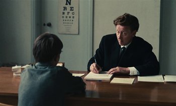 Movie still from “Kes” (1969), directed by Ken Loach – A man sitting at a table writing on a piece of paper; Medium shot, Over the shoulder angle