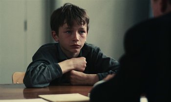 Movie still from “Kes” (1969), directed by Ken Loach – A young boy sitting at a table with his hands crossed; Close Up shot, Over the shoulder angle