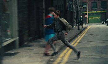 Movie still from “Kes” (1969), directed by Ken Loach – A man running down the street with a backpack on his back; Wide shot, High angle