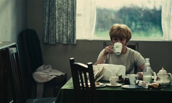 Movie still from “Kes” (1969), directed by Ken Loach – A woman sitting at a table drinking from a cup; Medium shot, Over the shoulder angle