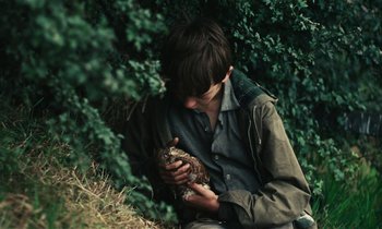 Movie still from “Kes” (1969), directed by Ken Loach – A man sitting on the ground with a bird in his hand; Close Up shot, Low angle