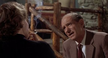 Movie still from “Kid Galahad” (1962), directed by Phil Karlson – An older man smoking a cigarette in front of a woman; Medium shot, Over the shoulder angle