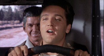 Movie still from “Kid Galahad” (1962), directed by Phil Karlson – A man driving a car while another man watches from behind him; Close Up shot, Low angle