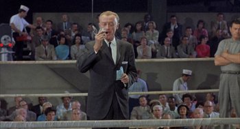Movie still from “Kid Galahad” (1962), directed by Phil Karlson – An older man in a suit and tie talking on a cell phone in front of an audience; Medium shot, High angle