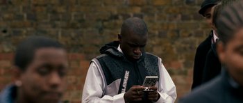 Movie still from “Kidulthood” (2006), directed by Menhaj Huda – A man is looking at his cell phone while standing in front of a brick wall; Close Up shot, High angle
