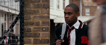 Movie still from “Kidulthood” (2006), directed by Menhaj Huda – A man in a suit and tie standing in front of a brick wall; Close Up shot, Over the shoulder angle