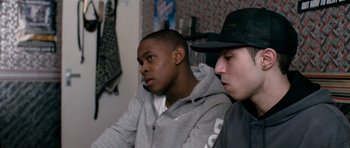 Movie still from “Kidulthood” (2006), directed by Menhaj Huda – Two young men sitting next to each other in a room; Medium shot, Over the shoulder angle